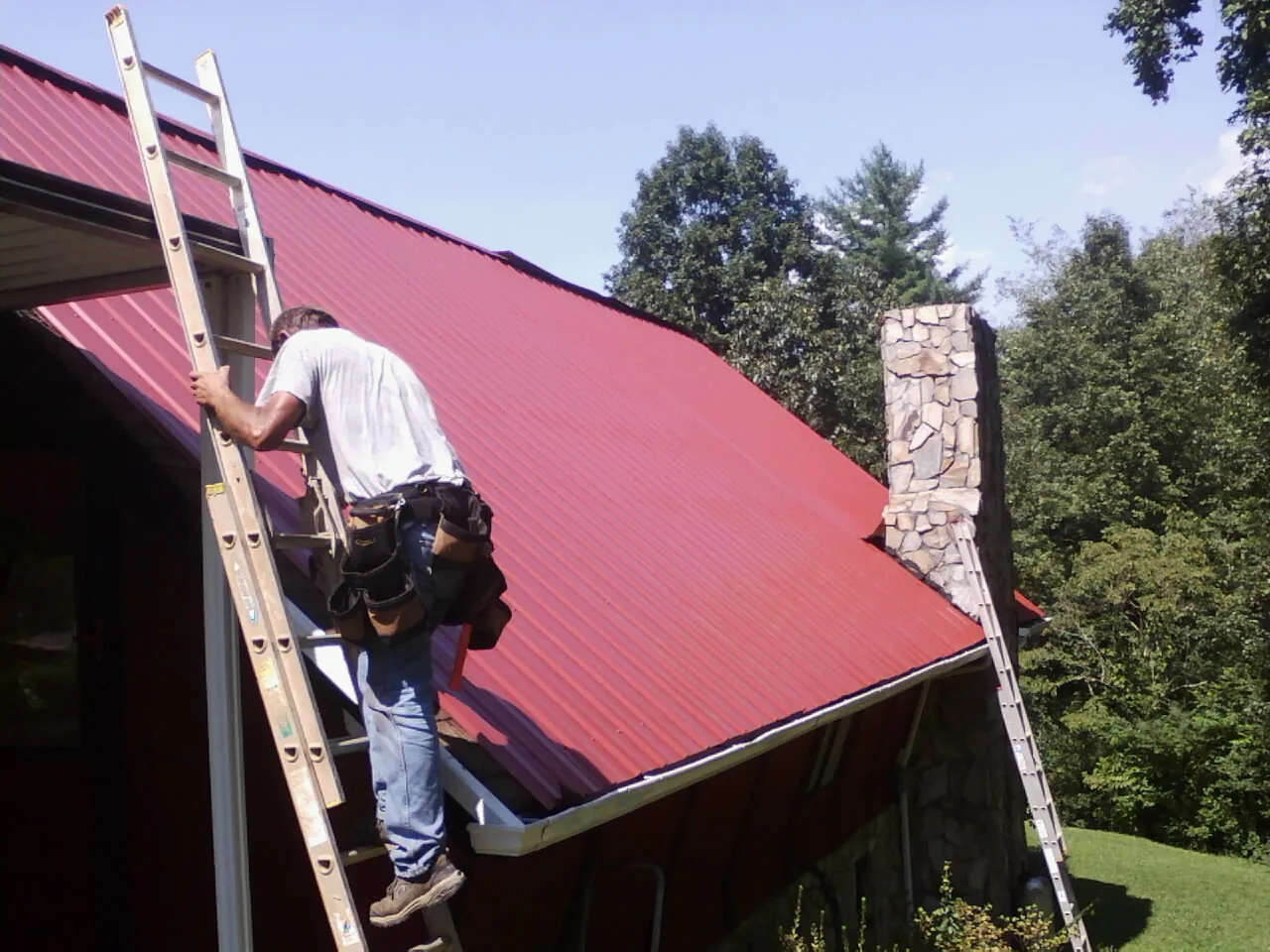 Roofer on a ladder beside a red metal roof and stone chimney — Soffit & Fascia Repair by Roofing Brothers in Mc Kees Rocks